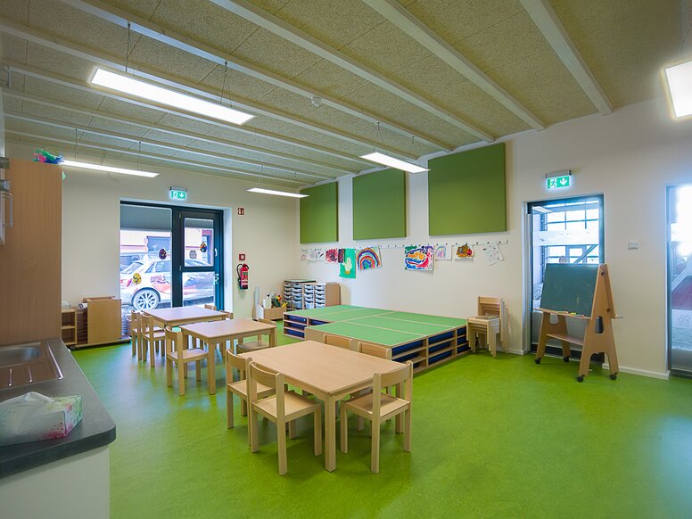Salle de groupe de la crèche "In der Kammgarnspinnerei" (dans la filature de laine peignée) Salle de groupe d'une crèche avec un sol vert et des tables et chaises en bois